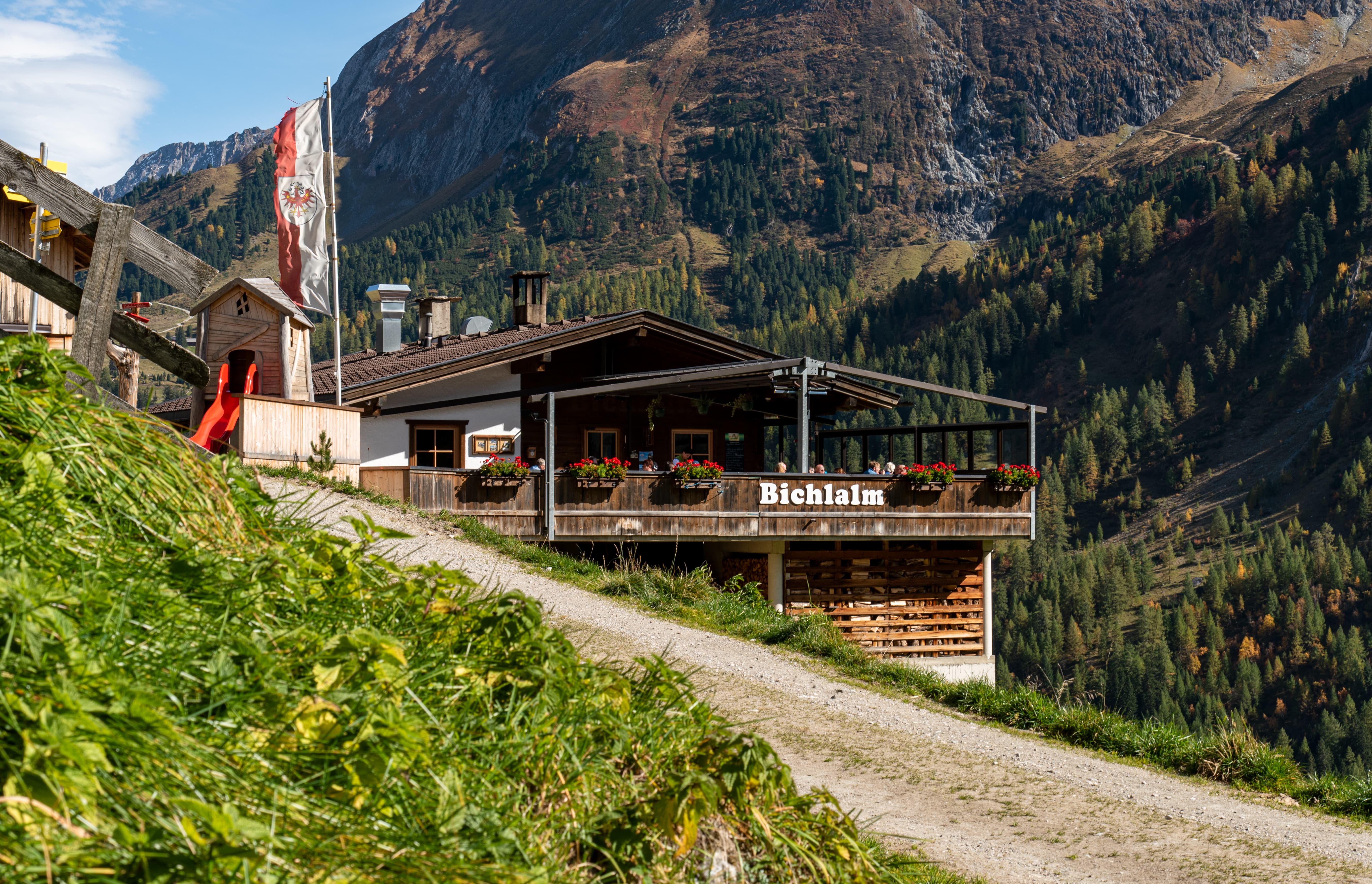 Berggasthaus Bichlalm mit Terrasse, Tiroler Flagge und Berglandschaft mit Nadelbäumen im Herbst.