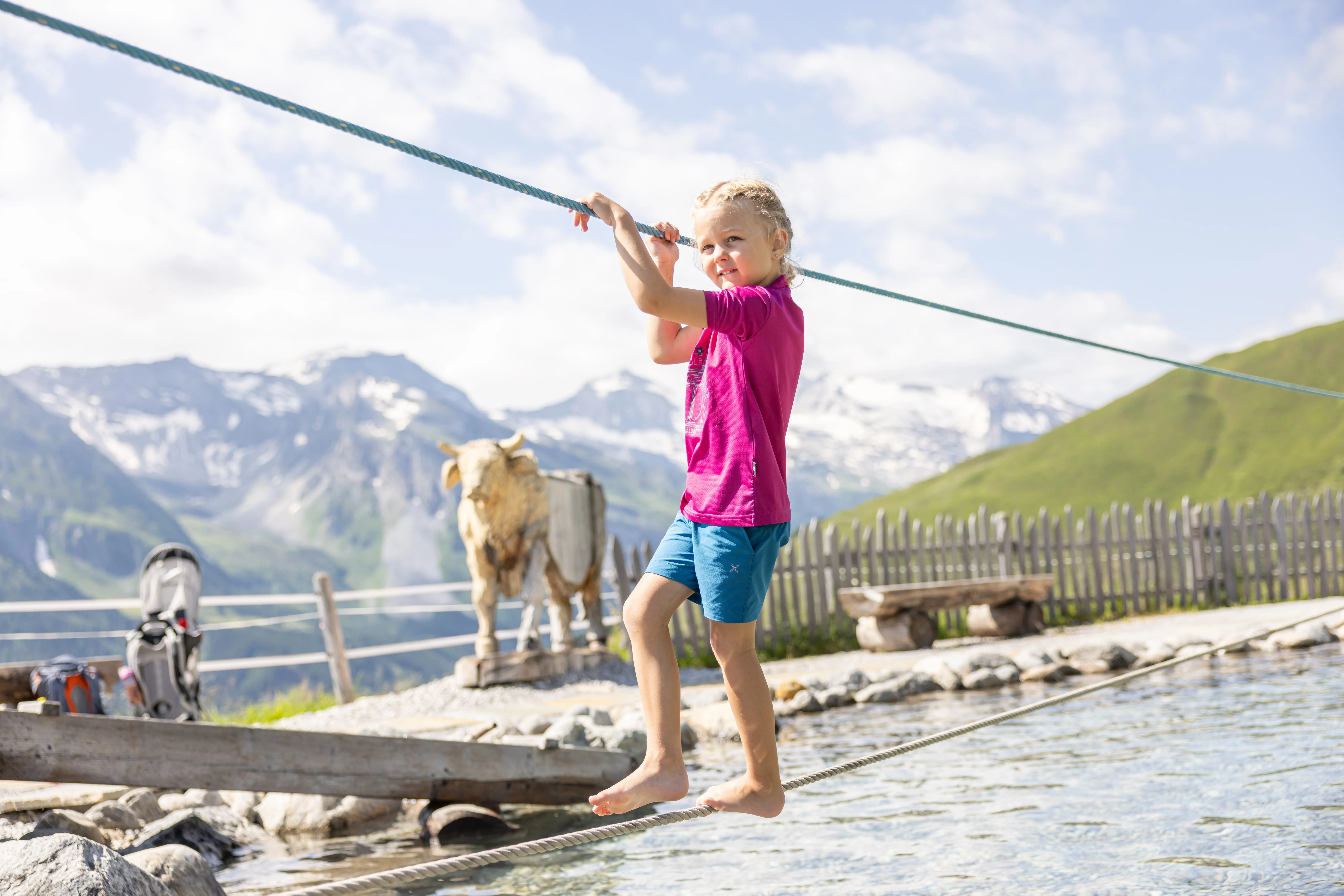 Kind balanciert barfuß auf einem Seil über Wasser vor Bergkulisse und Holzskulptur einer Kuh.