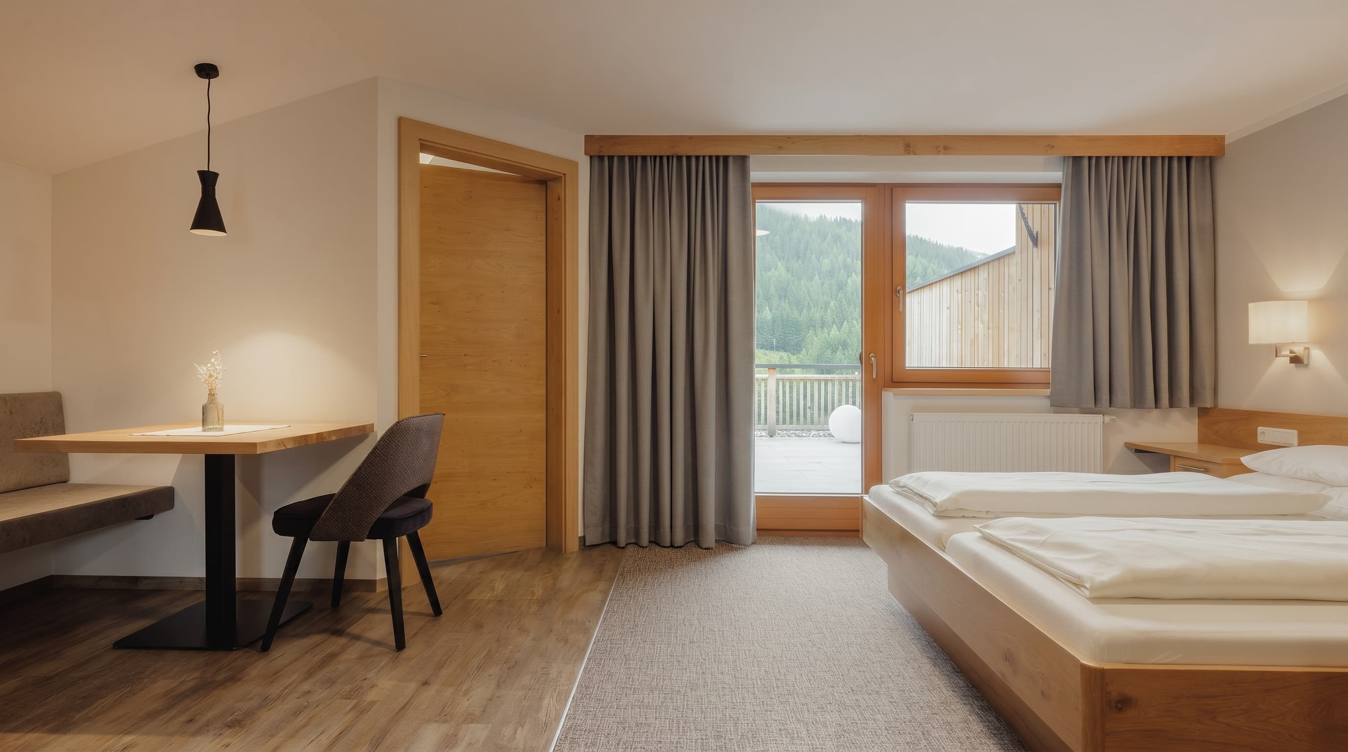 Helles Hotelzimmer mit Holzmöbeln, Doppelbett, Tisch, Stuhl und Blick auf bewaldete Berge.