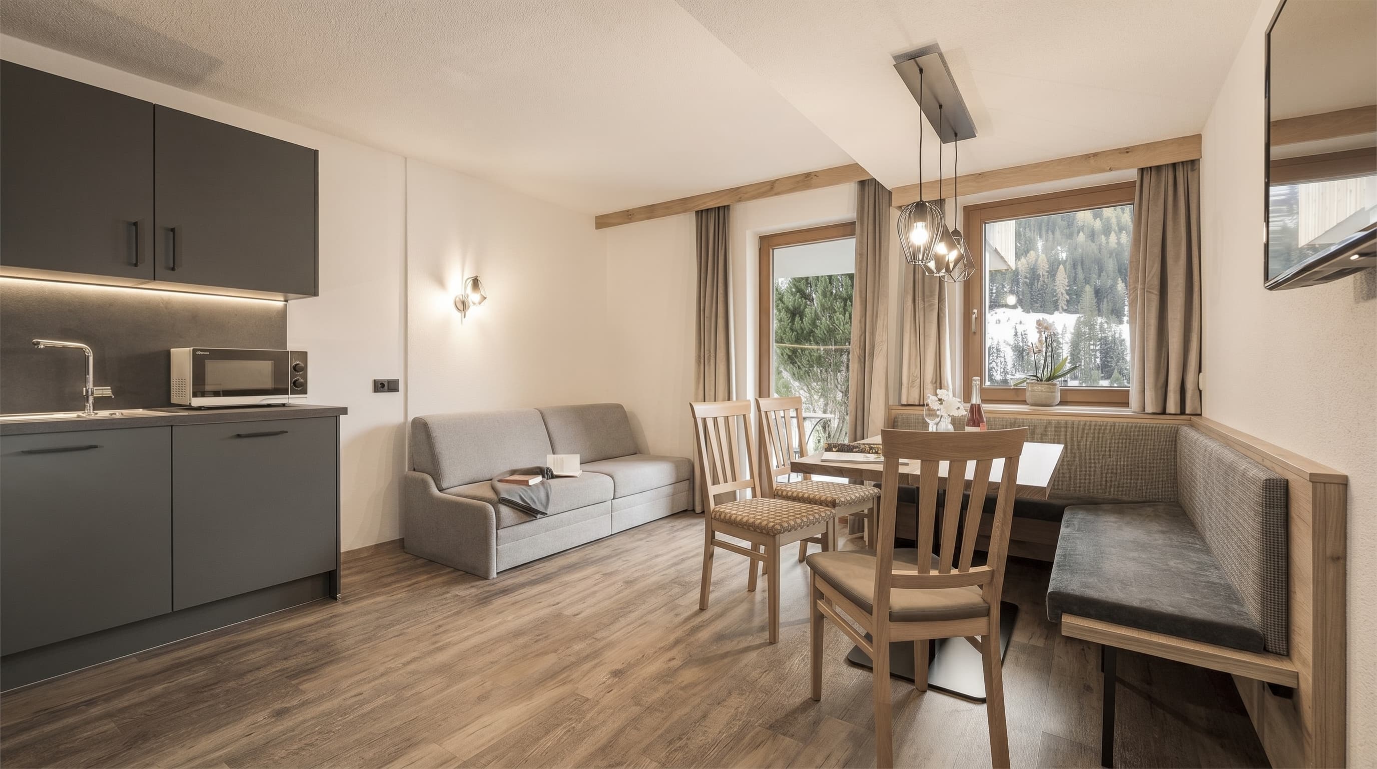 Helles Wohnzimmer mit grauer Couch, Holzstühlen, Eckbank, Esstisch und moderner Küche.