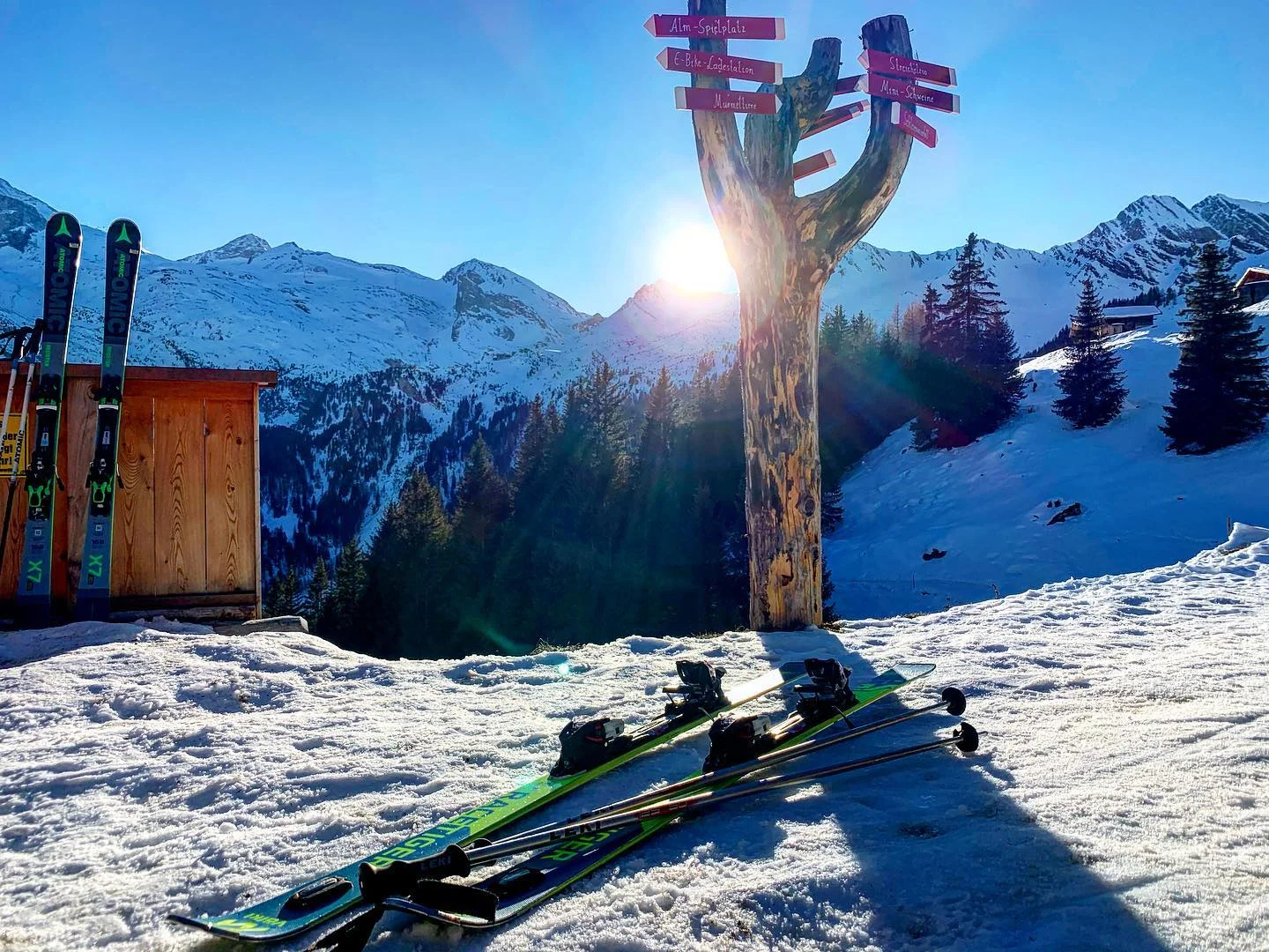 Skier gear laid on snow near wooden signpost and skis against cabin with snowy mountains and sun in background.
