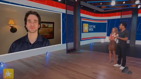 Two presenters on a studio stage watching a large screen showing a man speaking, with a sign reading '3 of the Week' in the background.
