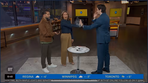 Three TV hosts standing around a small round table on a news set, two hosts smiling and one host with a raised hand.