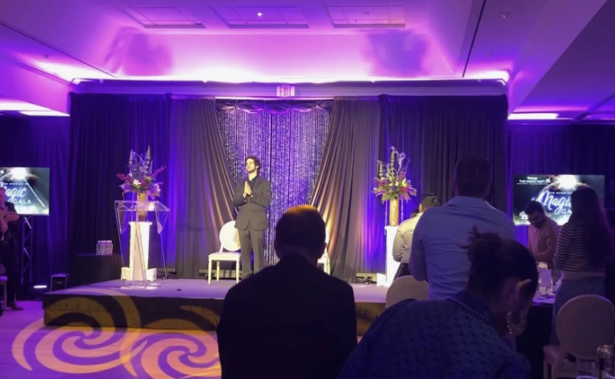 Man standing on a purple-lit stage with black curtains and floral arrangements, facing an audience seated at tables.