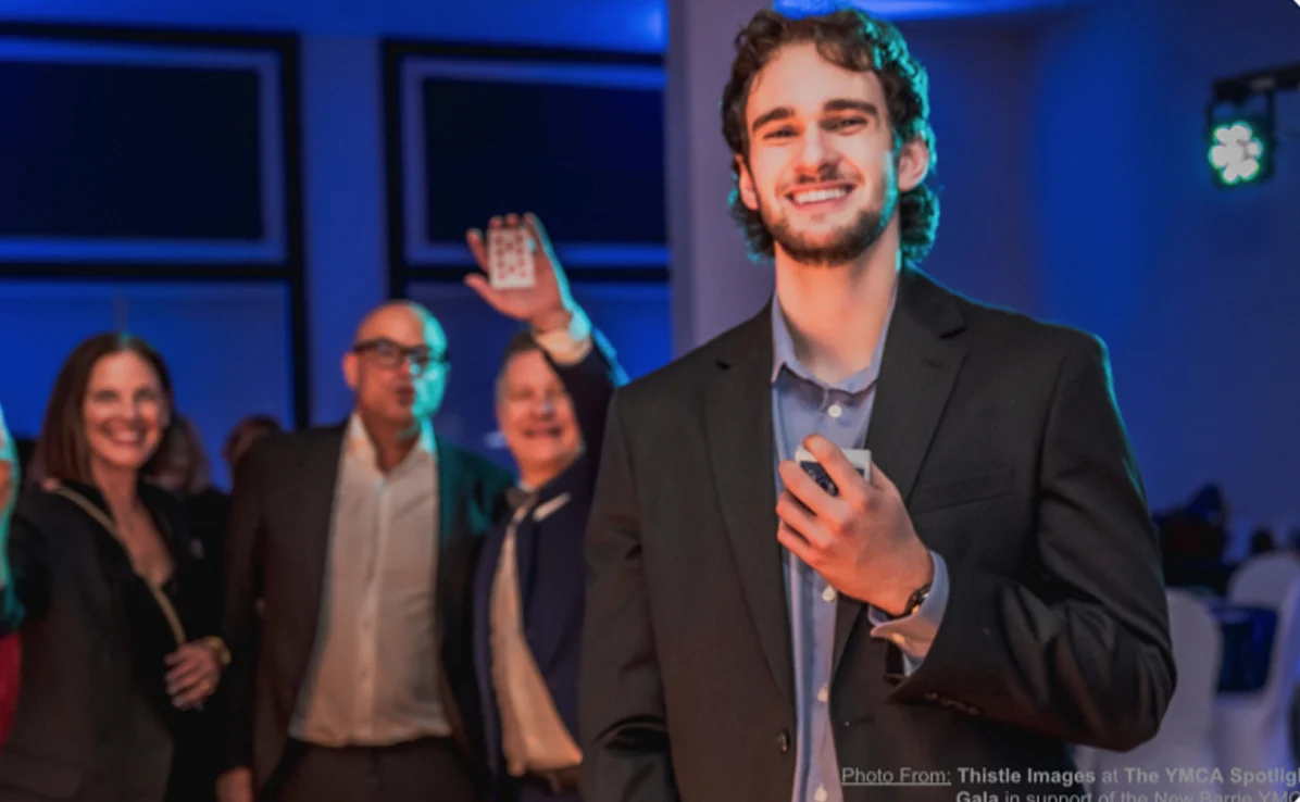 Smiling young man in a suit holding playing cards, with a group of people in formal attire in the background at an indoor event.