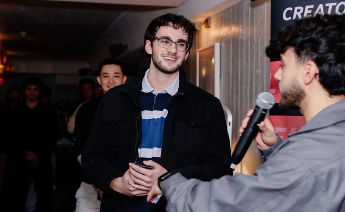Young man with glasses and curly hair smiling and holding a phone while another person speaks into a microphone during an indoor event.