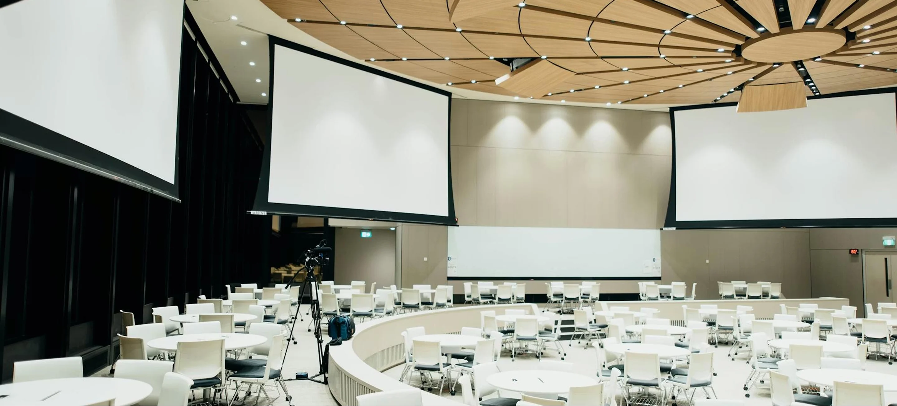Modern conference room with round tables, white chairs, three large blank projection screens, and a wooden ceiling design.