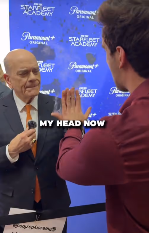 An older bald man in a suit and orange tie touching hands with a younger man in a maroon shirt, with a Starfleet Academy Paramount+ backdrop behind them and text overlay 'MY HEAD NOW'.