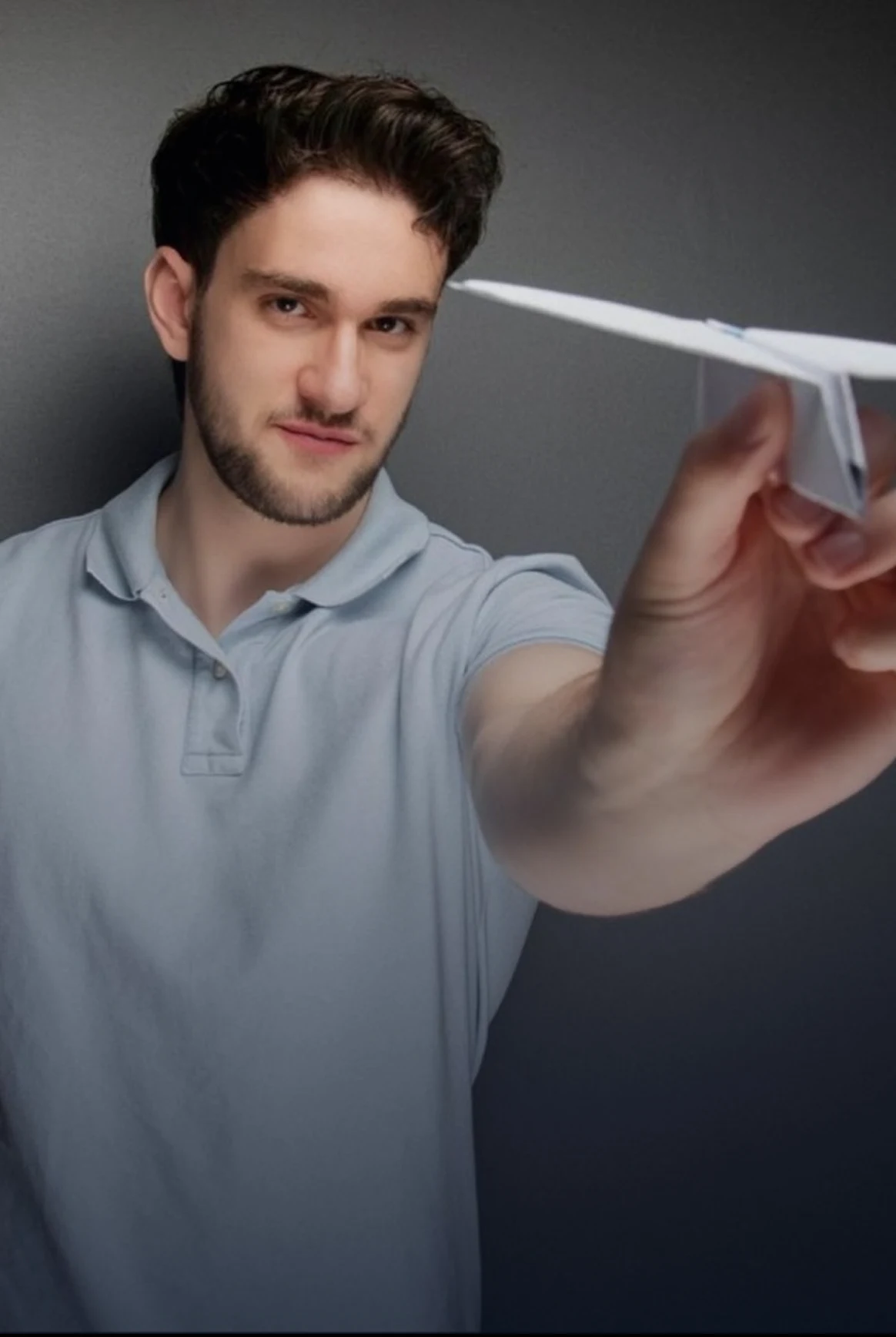 Young man with short dark hair and beard wearing a light blue polo shirt holding a white paper airplane aimed forward.