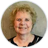 Portrait of a smiling older woman with short curly blonde hair wearing a black top and gray cardigan.