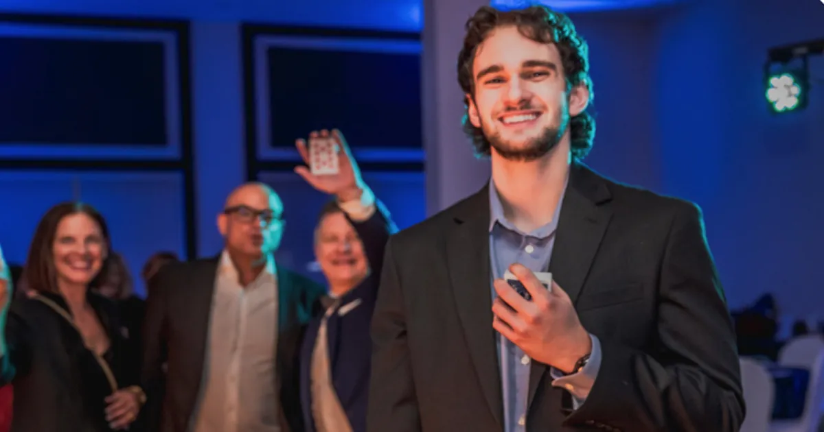 Young man smiling and holding playing cards with a group of people blurred in the background at a social event with blue lighting.