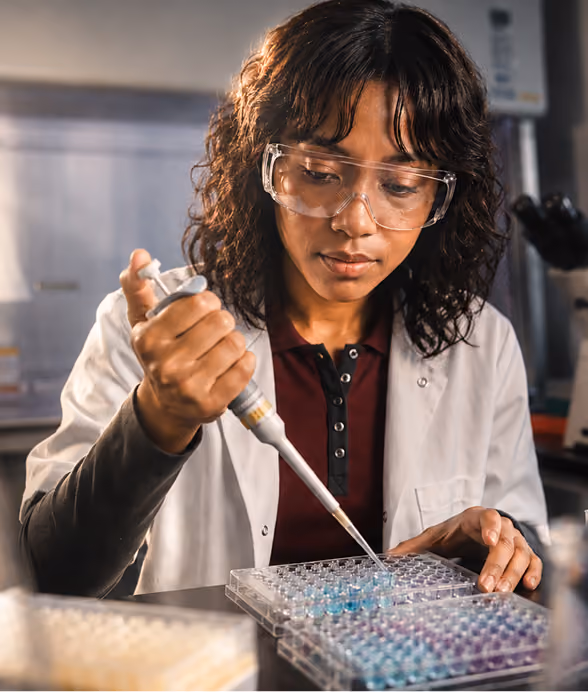 Scientist in gloves and lab coat using a pipette to extract liquid from a vial in a lab setting.