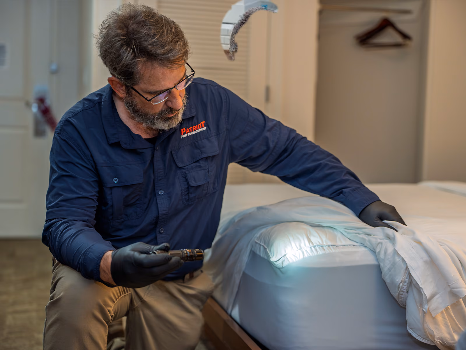 Pest control professional inspecting under bed covers with a flashlight in a hotel room