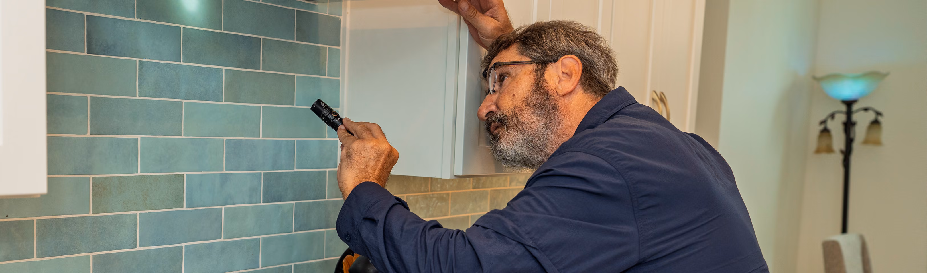 Man inspecting kitchen wall with blue tiles using a flashlight, checking near white cabinets.
