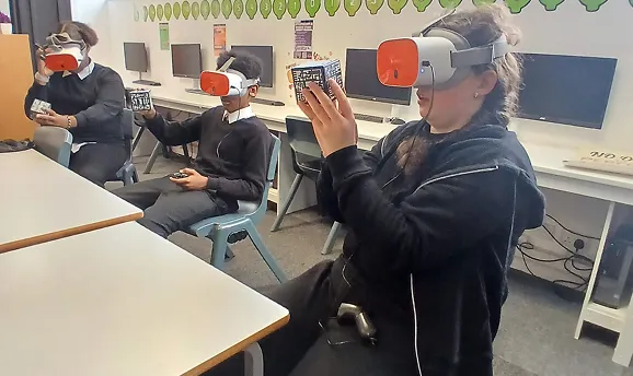 Three students sitting in a classroom using VR headsets and holding cube controllers.