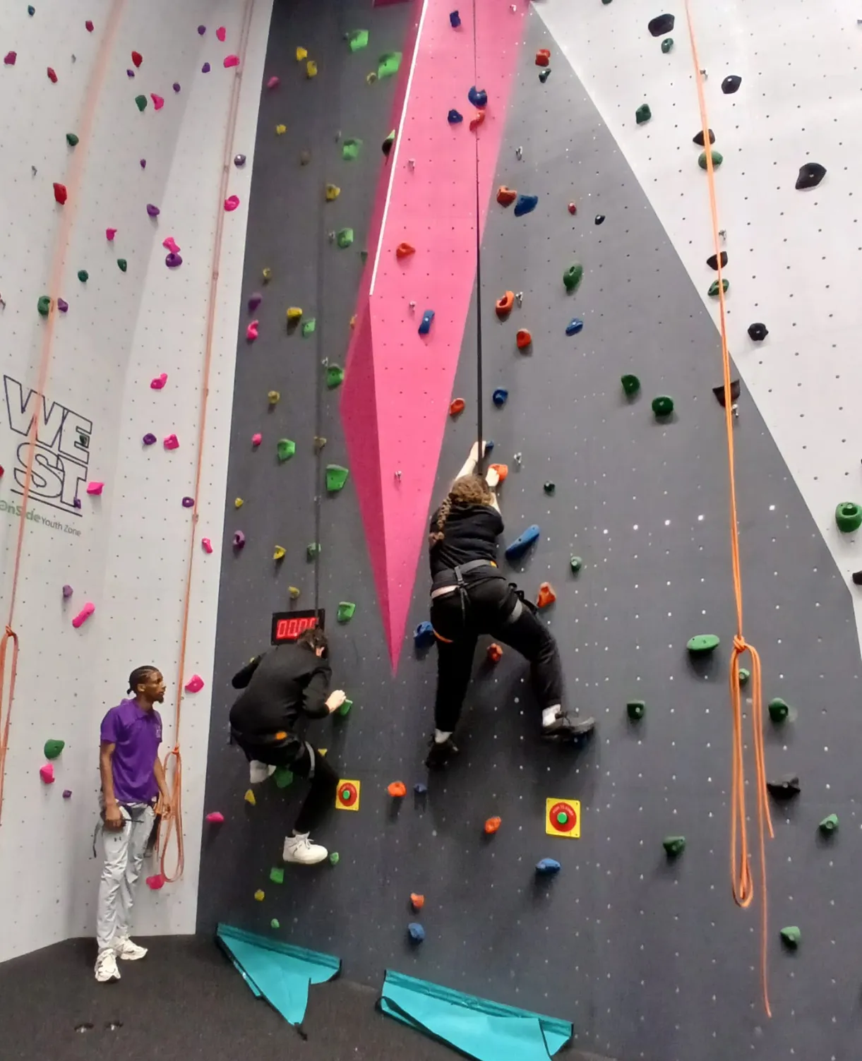 Two people climbing an indoor rock wall with a third person watching and holding the rope.