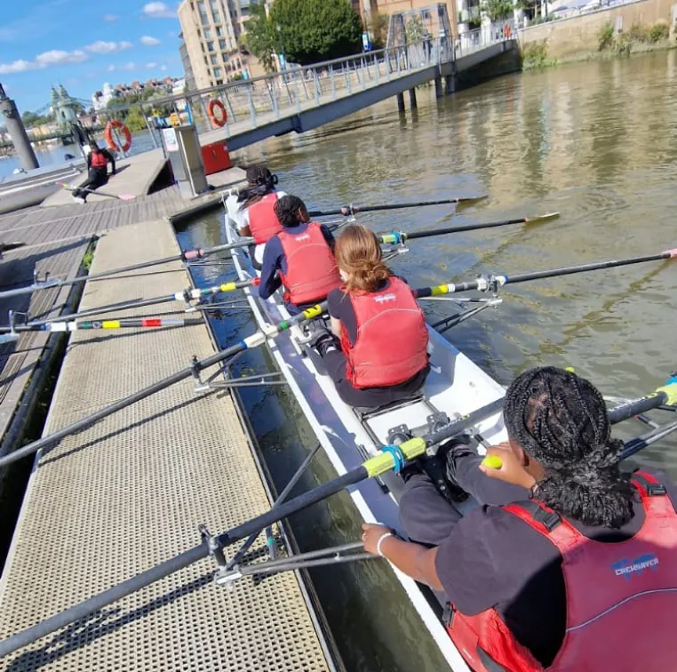 Four people in red life vests rowing together in a boat near a dock on a sunny day.
