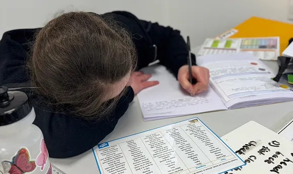 Person resting head on a table while writing in a notebook surrounded by papers and a water bottle.