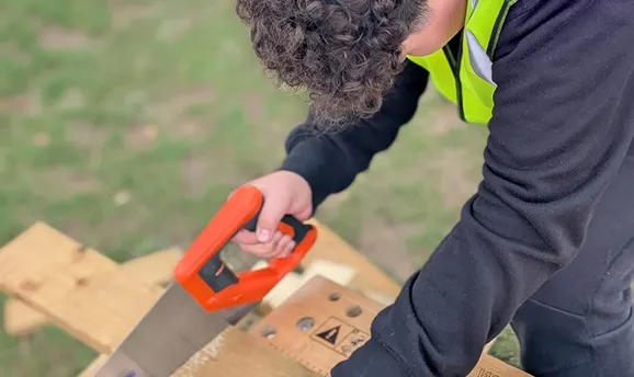 Child in safety vest using a hand saw to cut wooden boards outdoors.