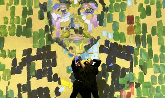 Two people in black posing with arms raised in front of a colorful, pixelated mosaic face artwork.