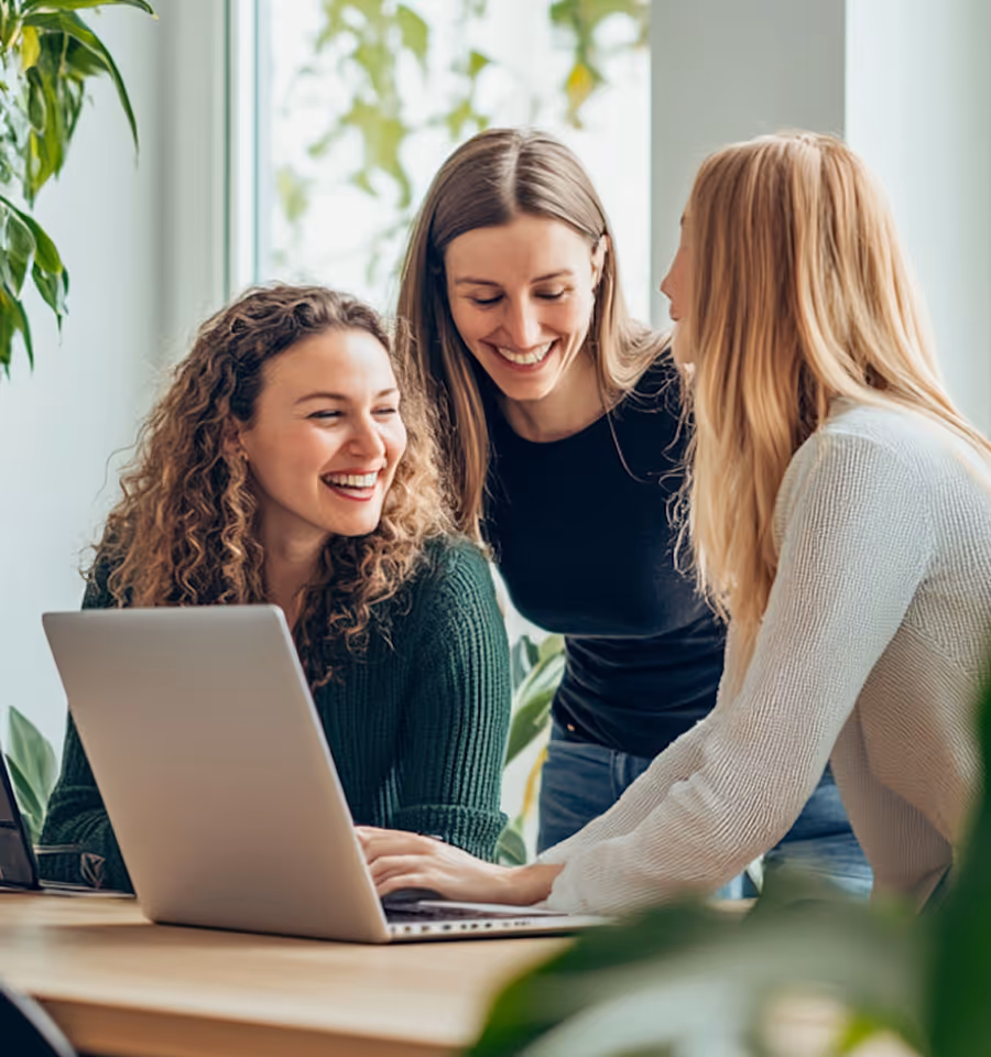 Three smiling women working together at a laptop in a bright, plant-filled office space.