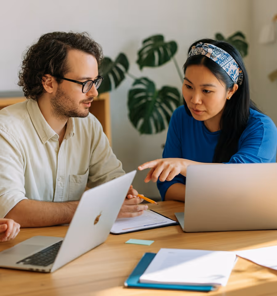 A man and woman discuss work at a table with laptops and documents in a bright office.