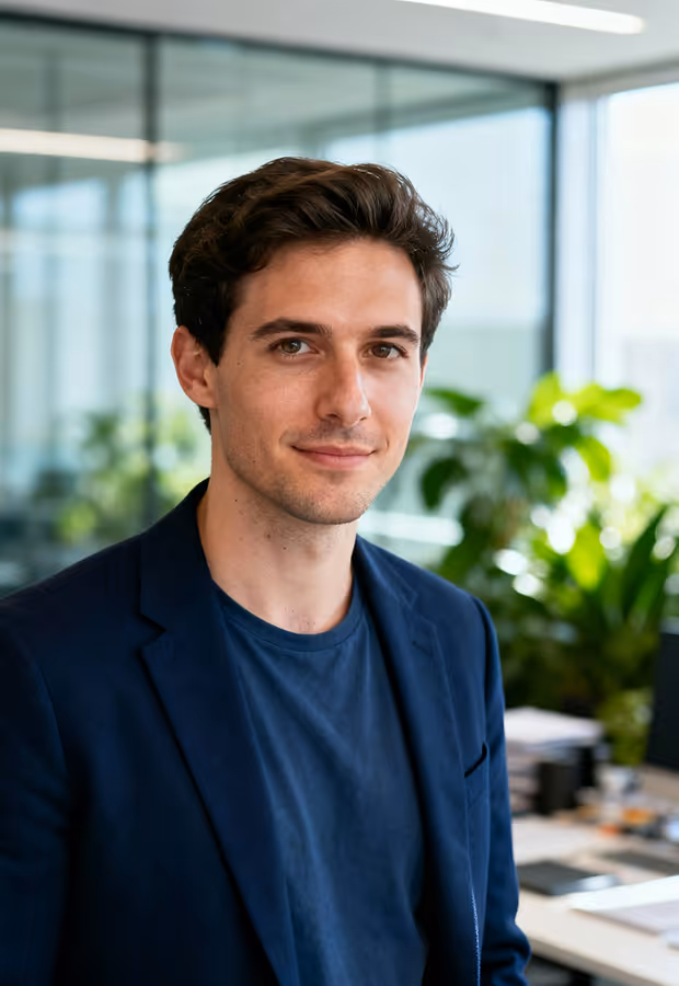 Young man in navy blazer and t-shirt smiling in a bright modern office with green plants.