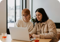 Two women sitting at a table working together on a laptop in a bright room.