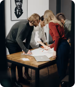 Four people standing around a wooden table, collaborating over documents and a laptop.