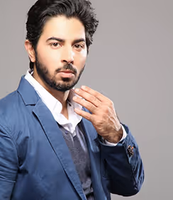 Man with dark hair and beard wearing a blue blazer and white shirt against a gray background.
