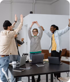 Three coworkers cheer and raise hands in an office with laptops on a table.