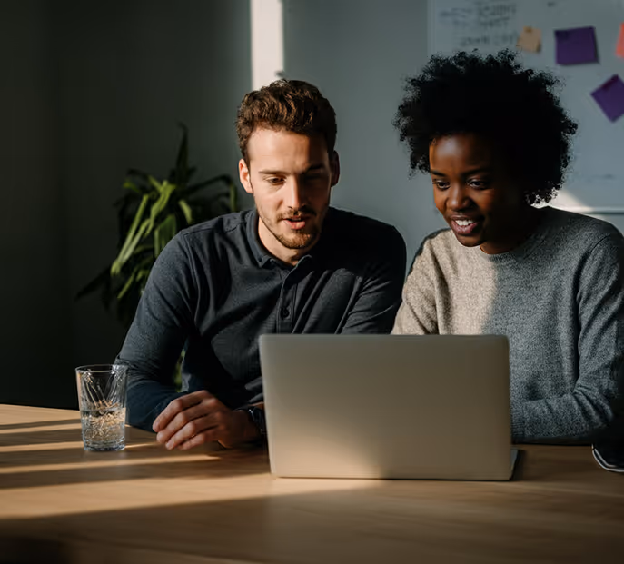 Man and woman sitting at a table, looking at a laptop together in a softly lit room.