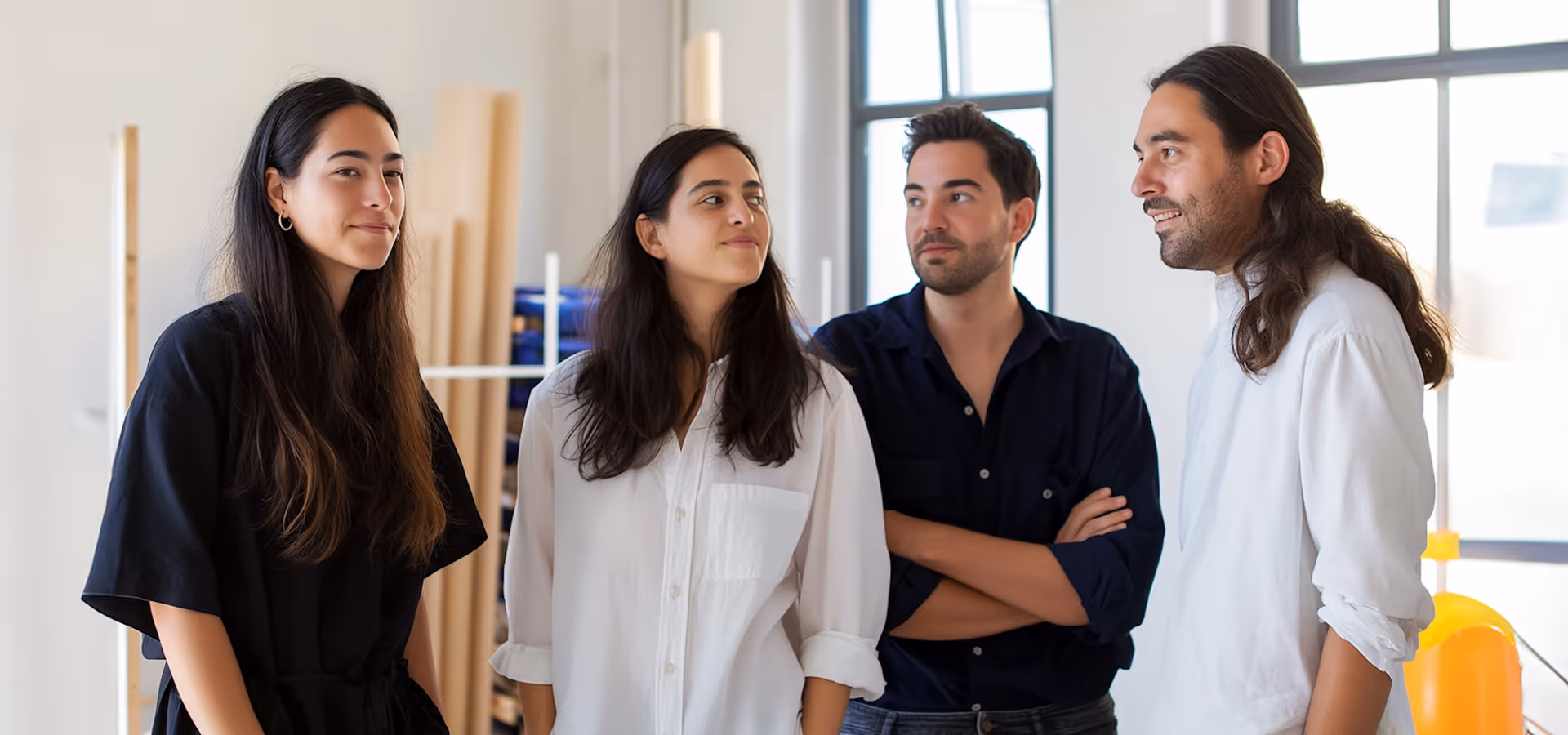 Four young adults standing indoors near large windows, two men and two women in casual shirts.