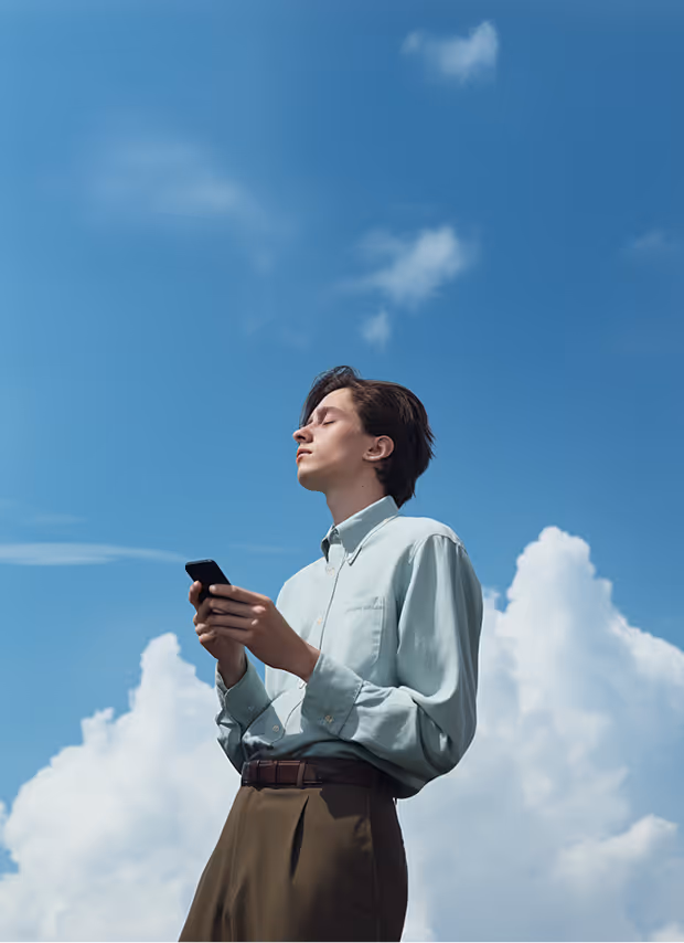 Young man with eyes closed holding phone and earbuds against a blue sky with clouds.