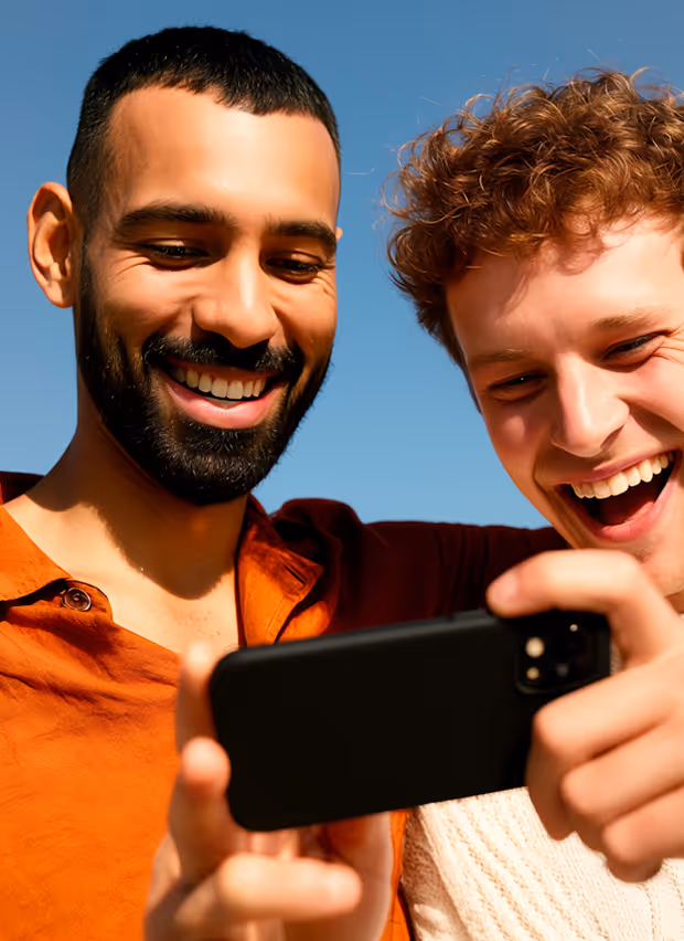 Two men smiling and looking at a smartphone against a clear blue sky.