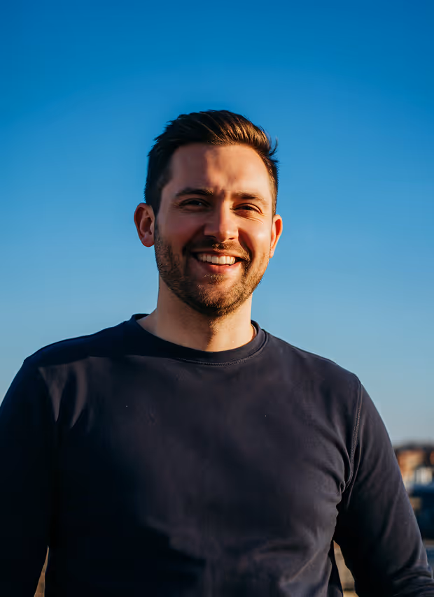 Smiling man with short hair and beard wearing a dark shirt against a clear blue sky.
