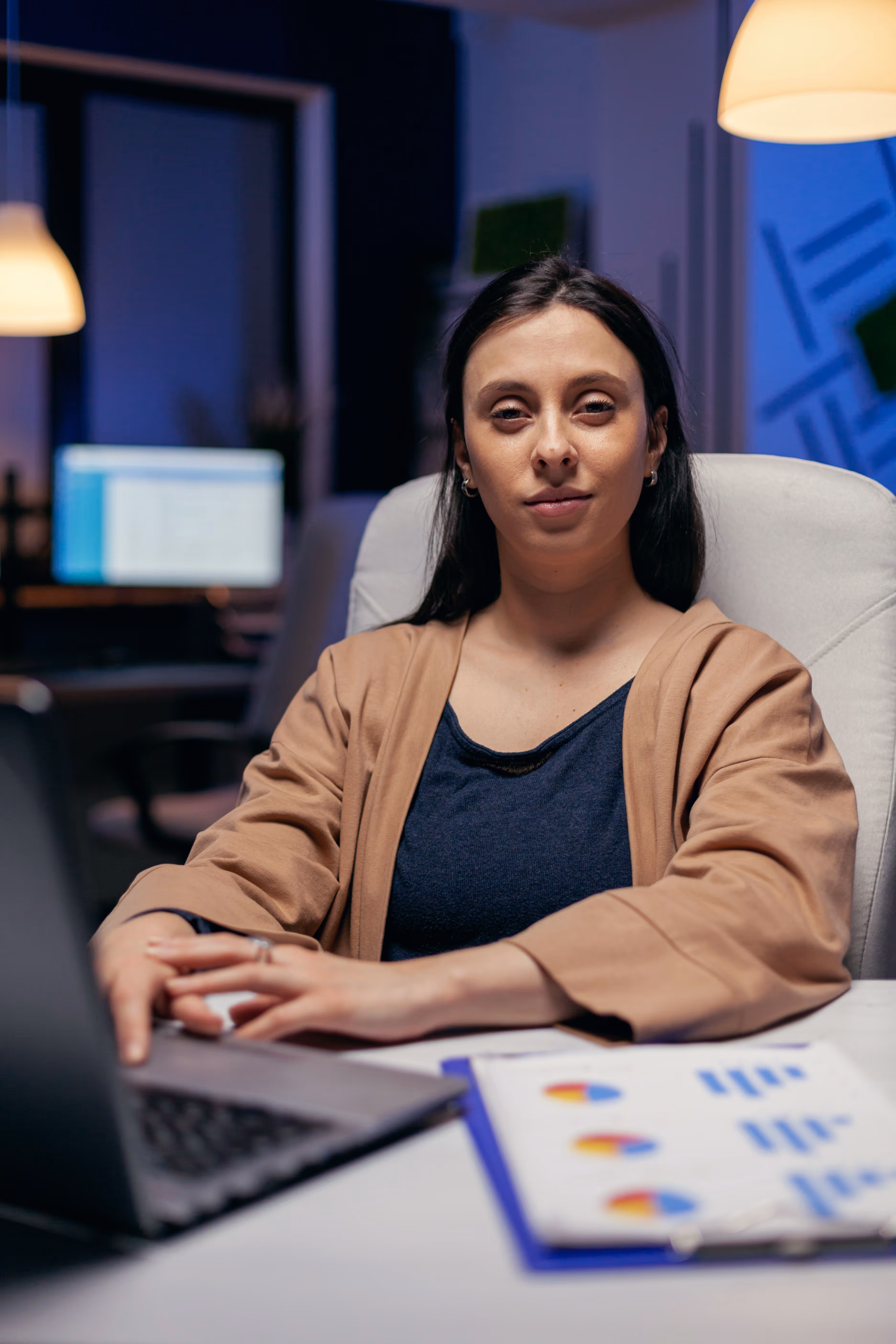 Woman sitting at desk with laptop and charts in office with dim lighting.
