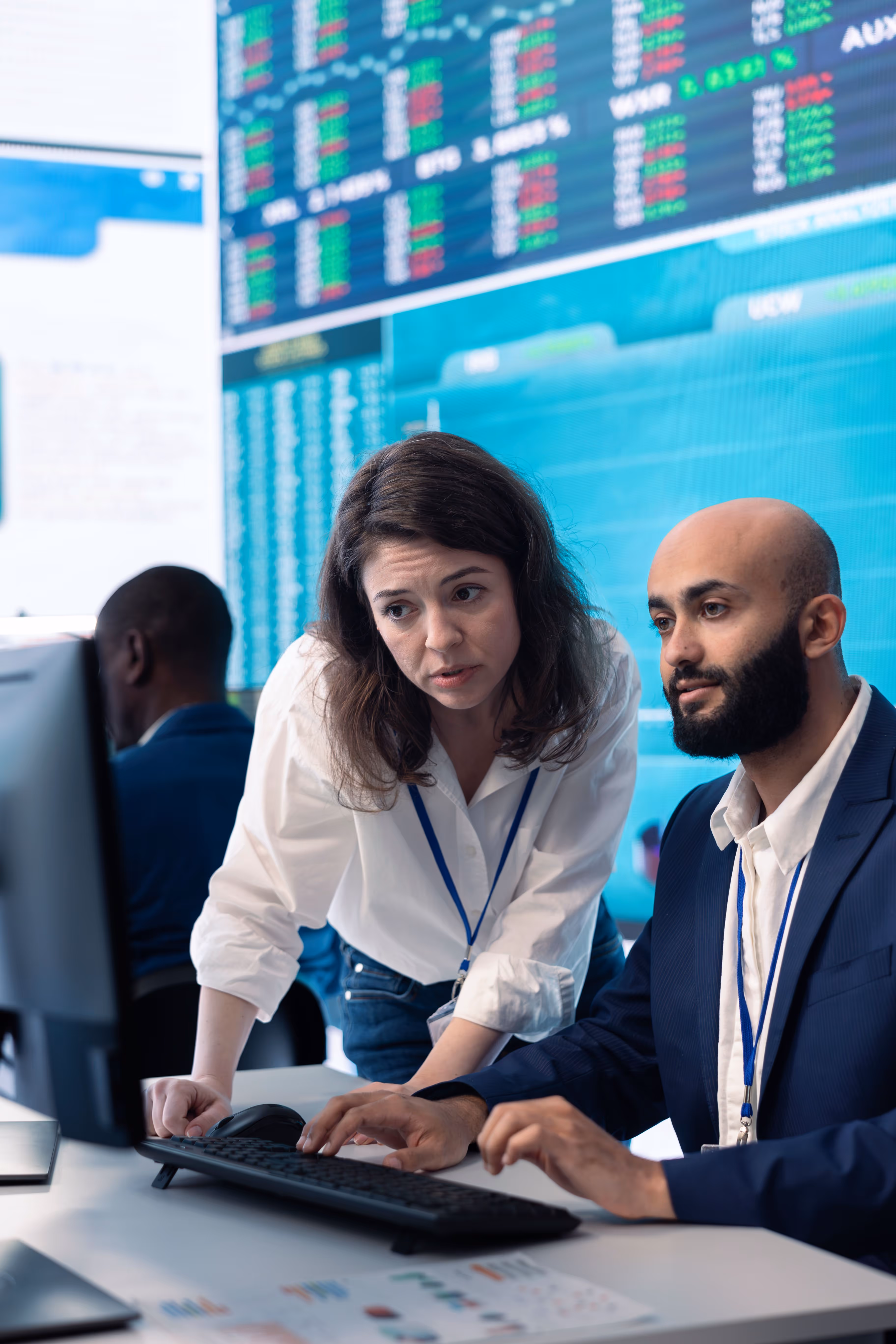 Colleagues analyzing data on a computer with stock market charts on large screens behind them.