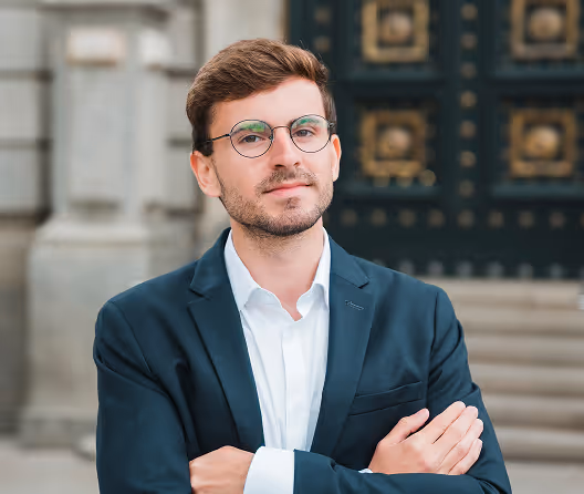 Confident young man with glasses in suit crossing arms outdoors near ornate door and stone wall.