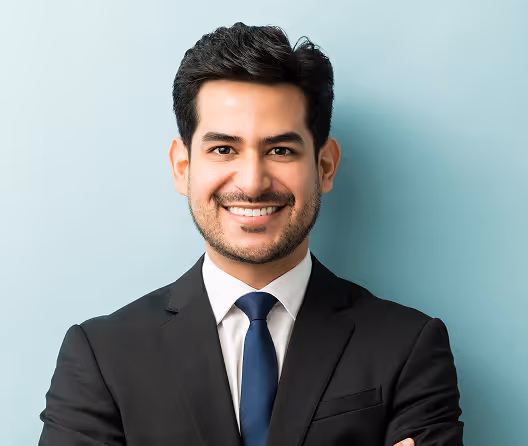 Smiling man in a black suit with white shirt and blue tie against a light blue background.