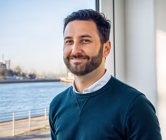 Smiling man with dark hair and beard wearing a green sweater by a waterfront window.