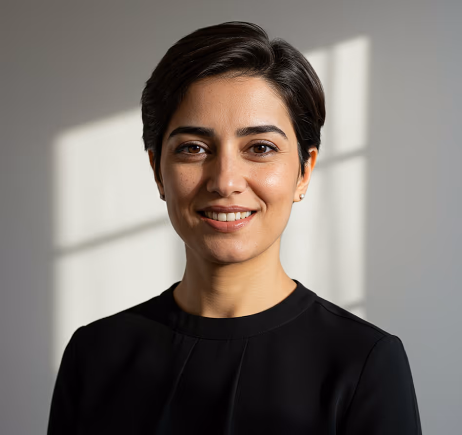 Smiling woman with short dark hair wearing a black top, with window shadows on a gray wall behind her.
