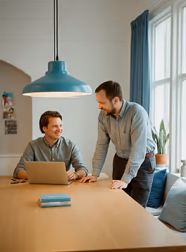 Two men smiling and discussing work at a table with a laptop under a blue hanging lamp.