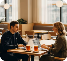 Man and woman sitting at a cafe table with drinks and a laptop, engaged in conversation.