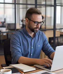 Man with glasses and beard working on laptop at desk with notebook and coffee cup.