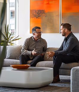Two men in business casual talking on a gray couch in a modern office lounge area.