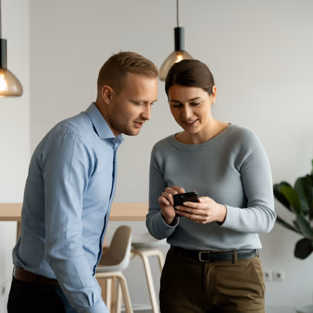 Two colleagues smiling and looking at a smartphone in a modern office setting.