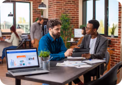 Two men discussing work at an office table with a laptop, documents, and a small plant.
