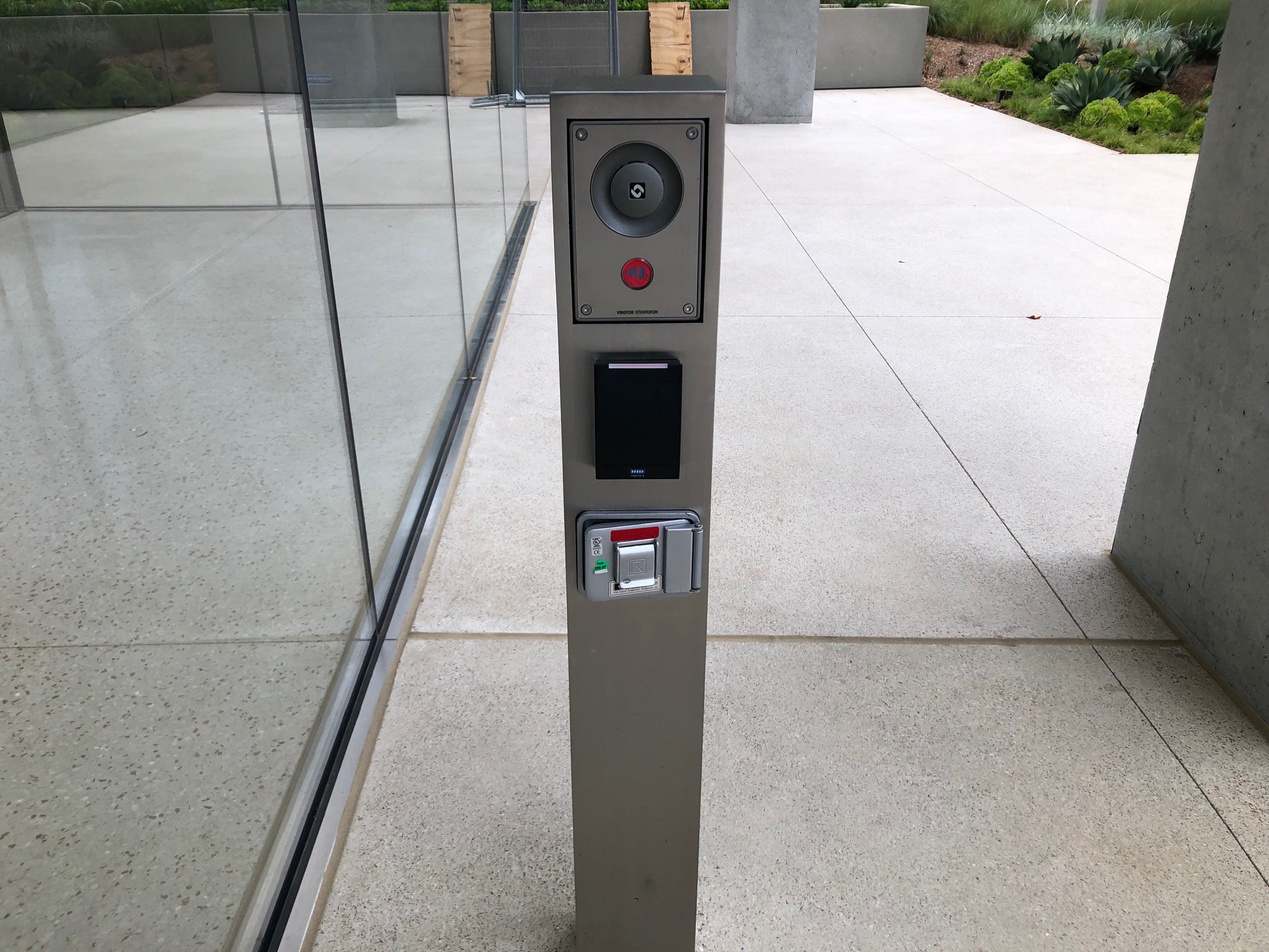 Modern intercom and access control panel on metallic pillar beside glass wall outdoors.