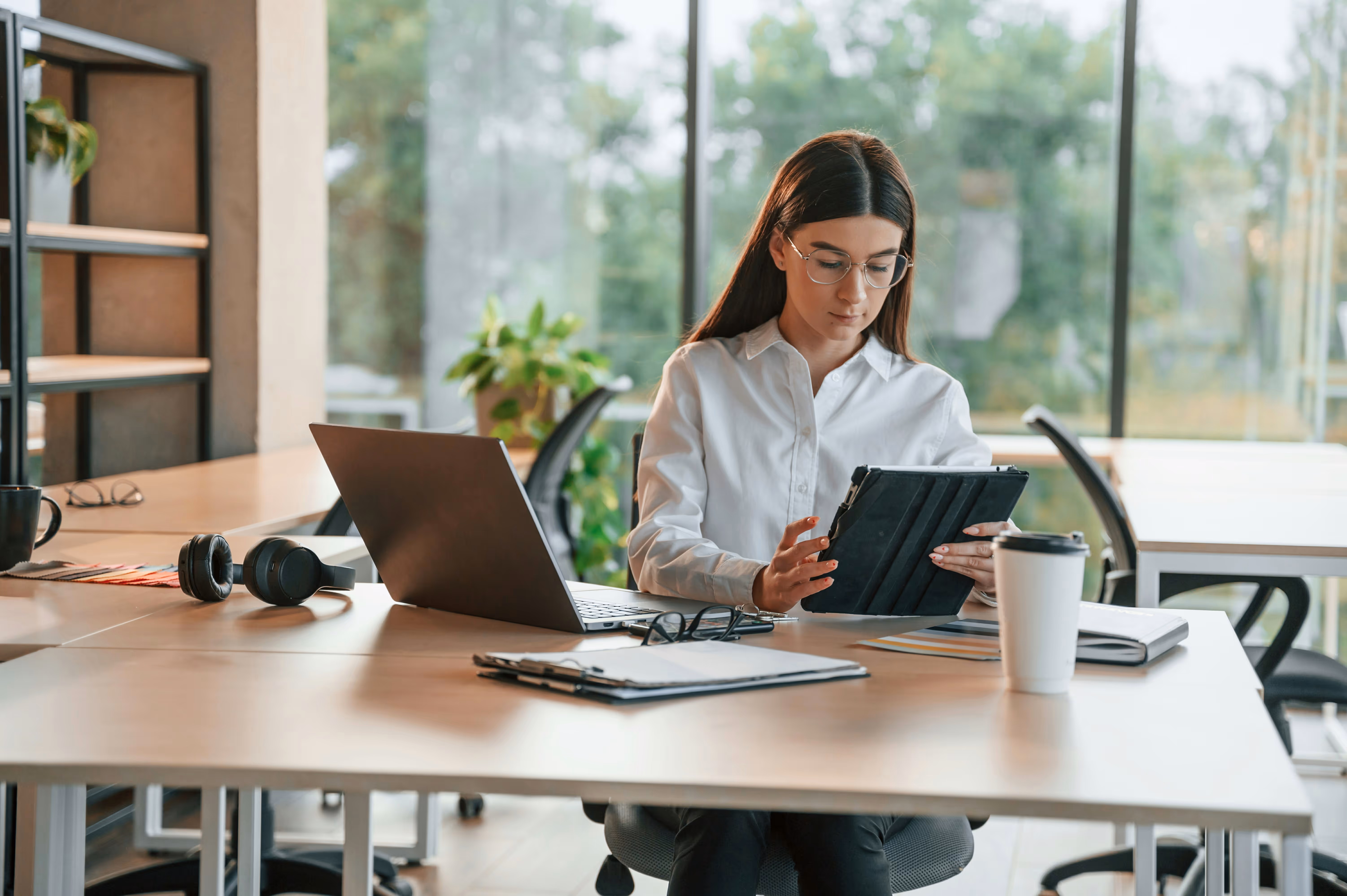 Femme en chemise blanche utilisant une tablette au bureau avec ordinateur portable, cahier et tasse à café.